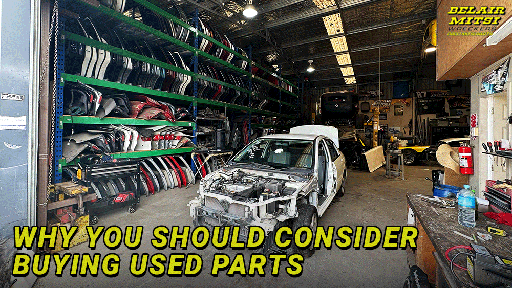 An indoor auto parts dismantling workshop with a partially stripped white Mitsubishi sedan in the center. Shelves line the back wall, displaying organized rows of various colored car panels, fenders, and doors.